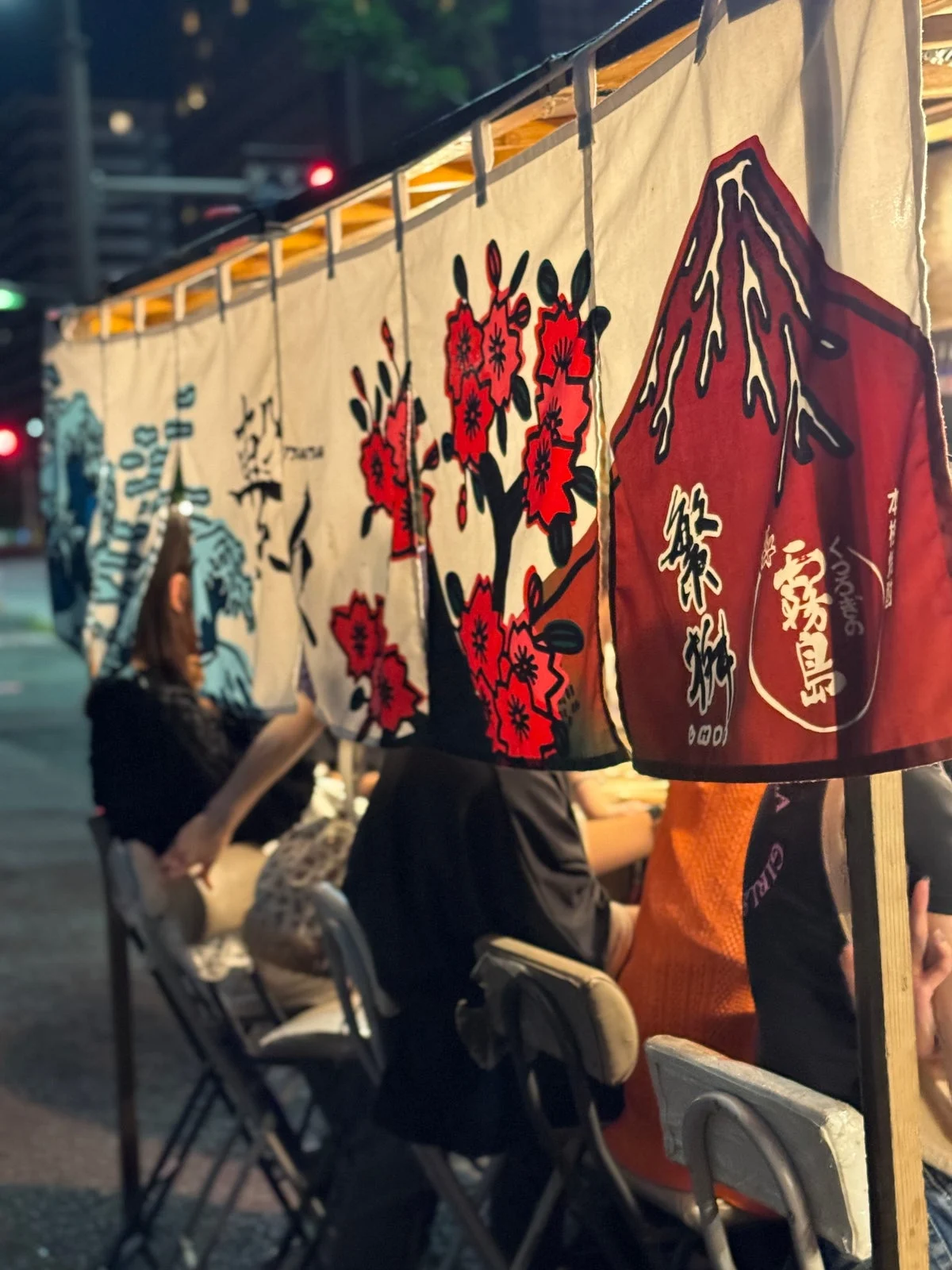 夜の中洲。赤い花模様ののれんがかかる屋台に並んで座る人たち。